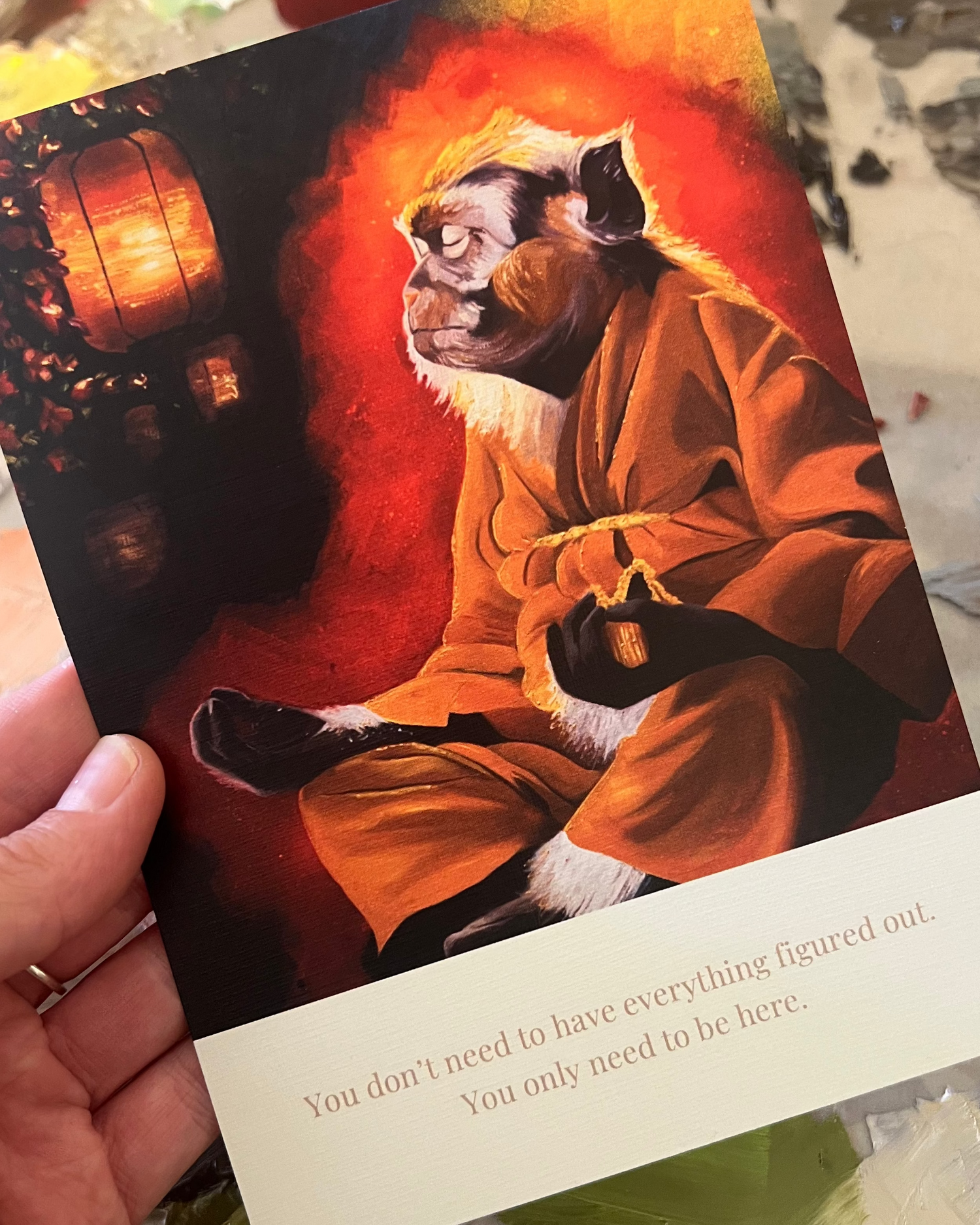 A hand holds a printed art card featuring a painted seated figure in orange robes against a fiery red and orange background, with a cream text band at the bottom. The card is photographed over a paint-splattered surface.