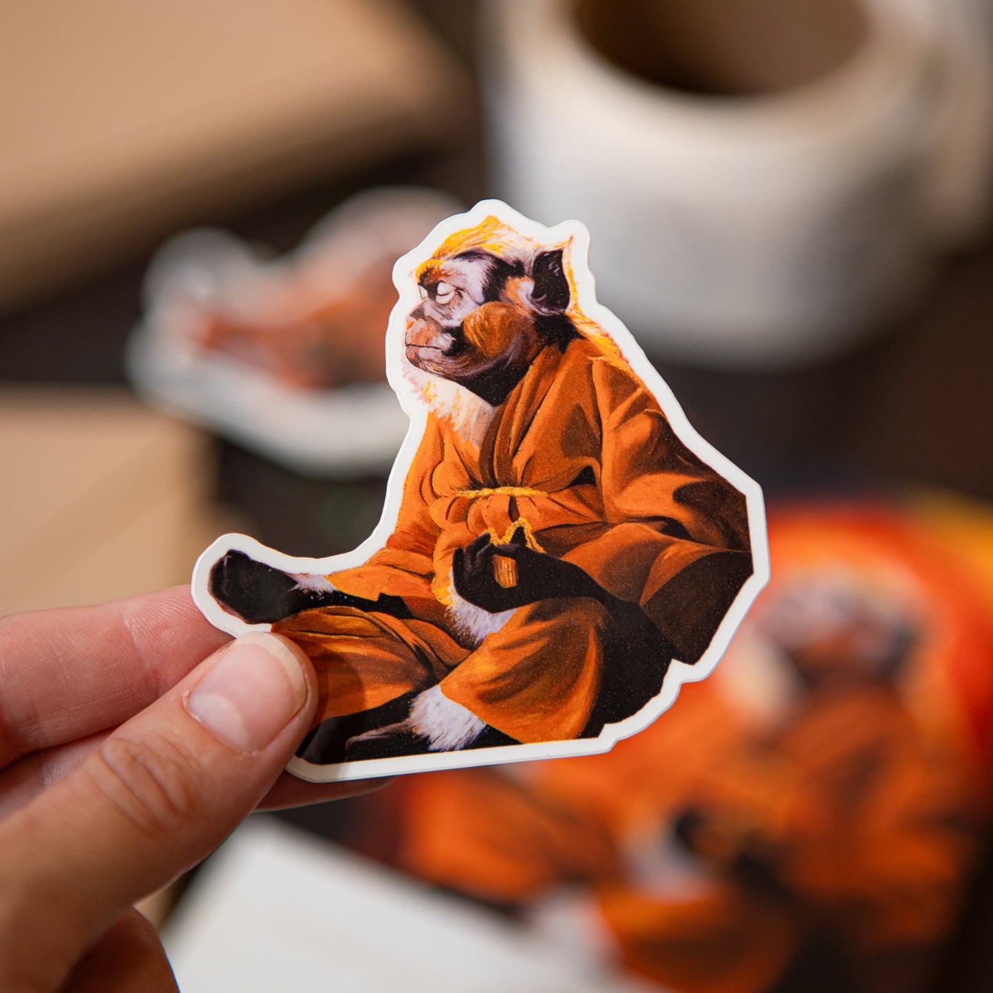 A hand holds a die-cut sticker featuring a monkey in an orange robe against a warm, blurred background with coffee cups and paper.