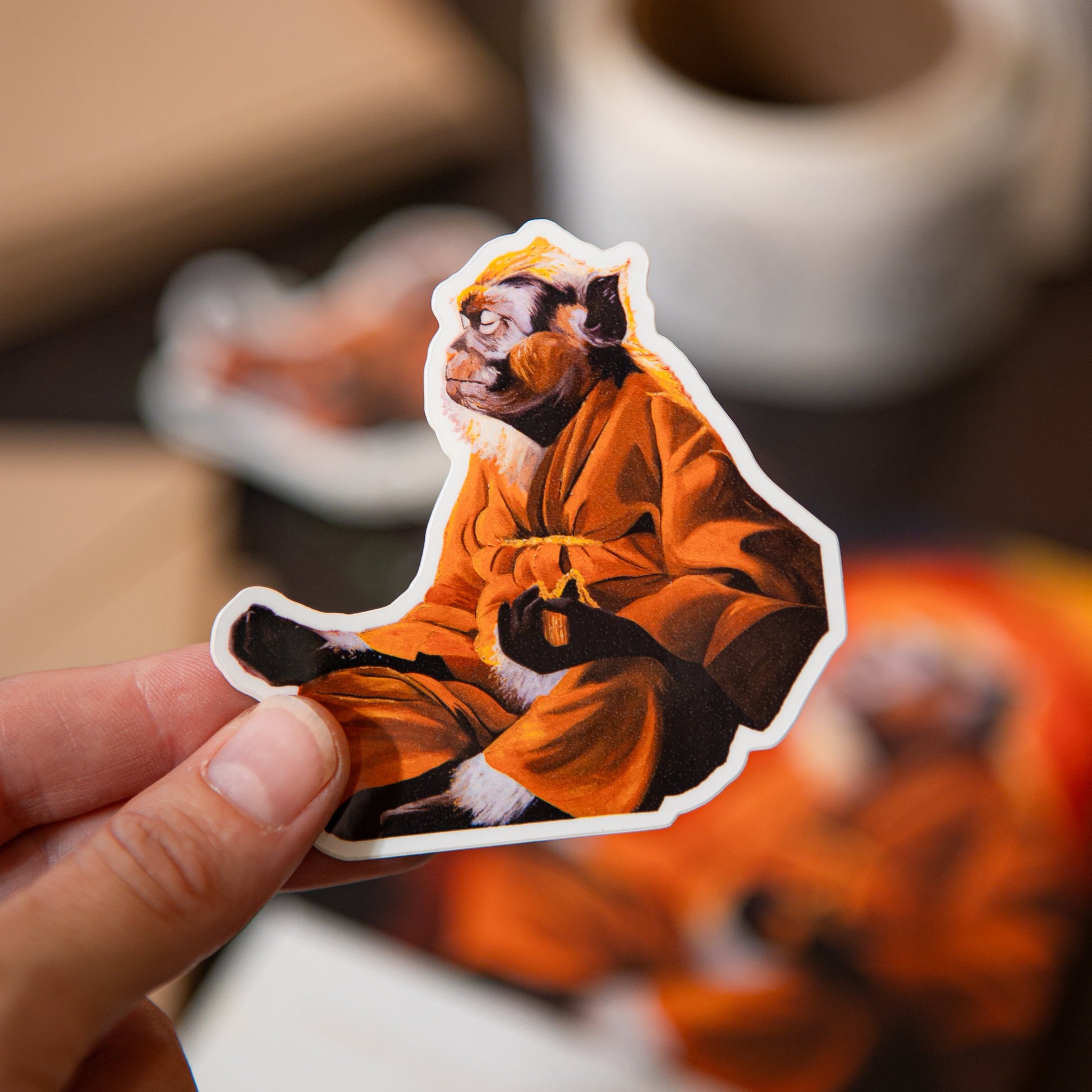 A hand holds a die-cut sticker featuring a monkey in an orange robe against a warm, blurred background with coffee cups and paper.